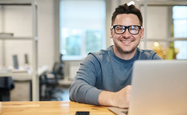 Happy man wearing glasses working remotely on laptop in modern office environment.